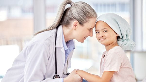Health care professional with a stethoscope sitting at eye level with a pediatric cancer patient, holding hands in a bright clinical setting.