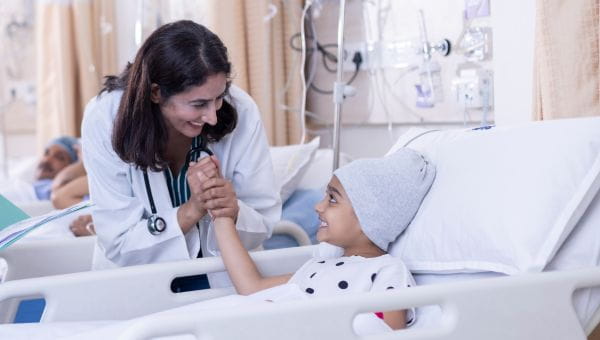 A physician holds the hand of a pediatric cancer patient who is lying in a hospital bed.