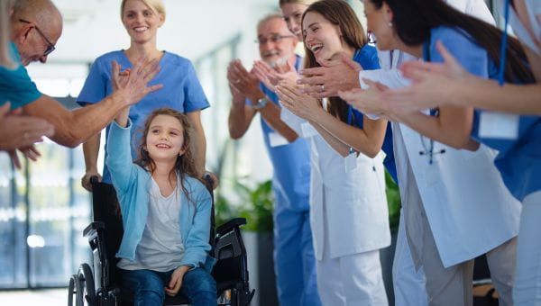 A pediatric patient in a wheelchair gives a high five to one health care professional while getting applause from a whole medical team.
