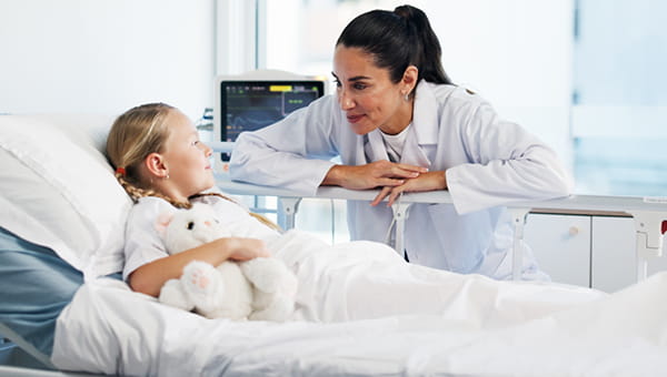 a female doctor bent down next to a hospital bed leaning towards a young girl with blond hair holding a stuffed animal, softly smiling at one another