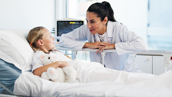 a female doctor bent down next to a hospital bed leaning towards a young girl with blond hair holding a stuffed animal, softly smiling at one another