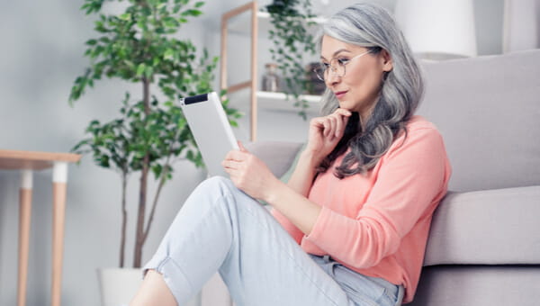 an elderly woman sitting against her couch and reading her tablet
