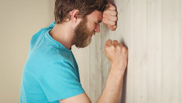 An anxious man rests his head on one hand against a wall, with the other fist clenched. 