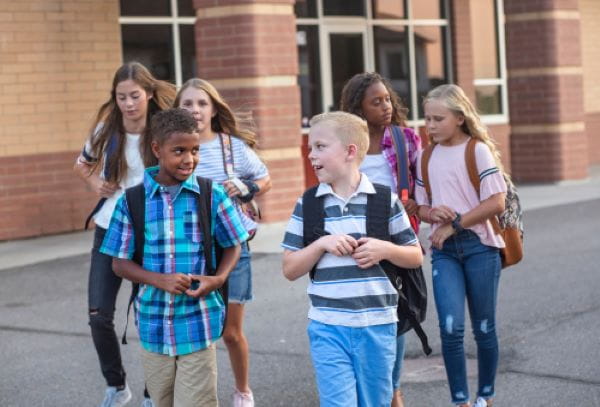 A diverse group of elementary aged kids walk away from a school and talk with each other.